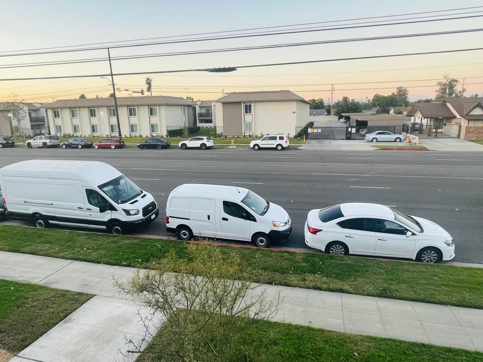 Three white vehicles parked along a roadside in front of residential buildings at dusk.